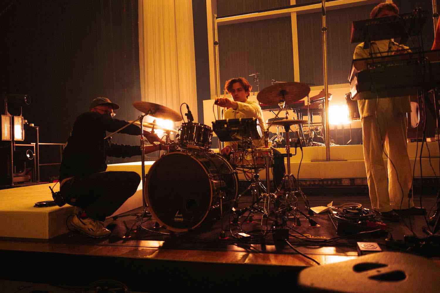 A drum kit and keyboard rig set up in a rehearsal room under warm lamps