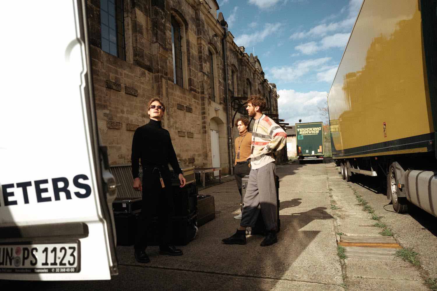 Two members of the band on a cobbled street beside a yellow truck and a stone building