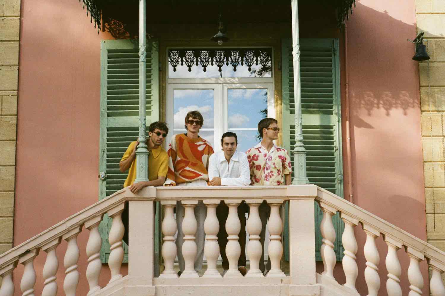 The four members of TORA leaning on the balcony railing of a pink building with green shutters