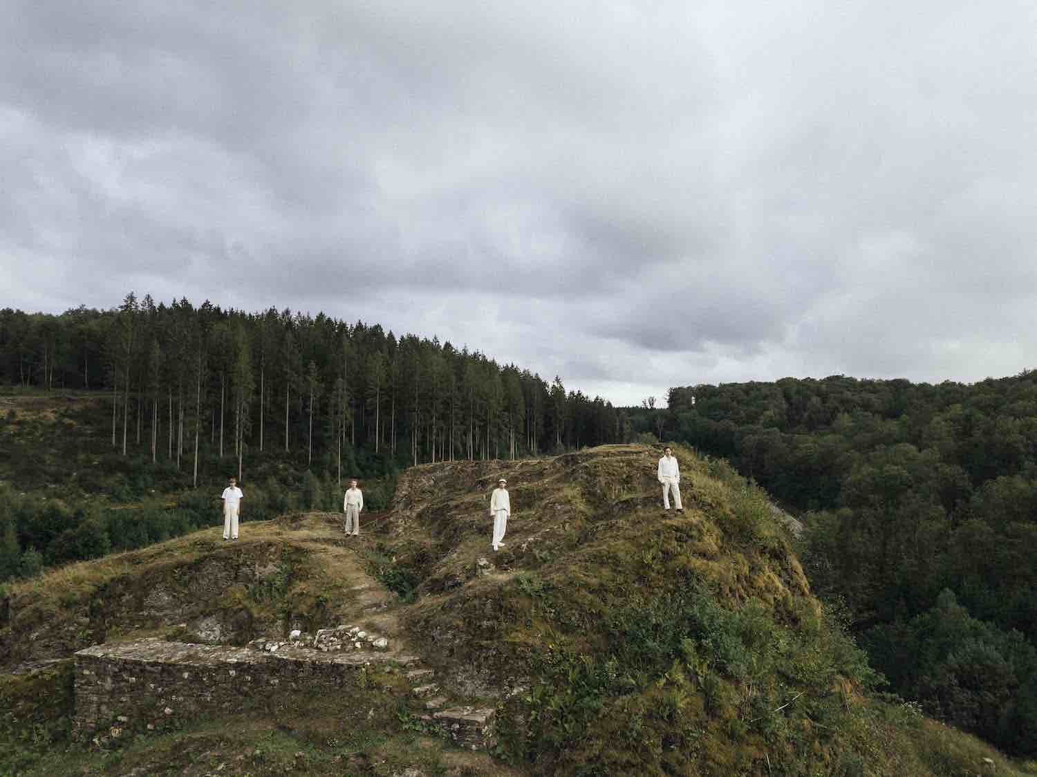 Four small figures dressed in white on a forested hilltop under an overcast sky
