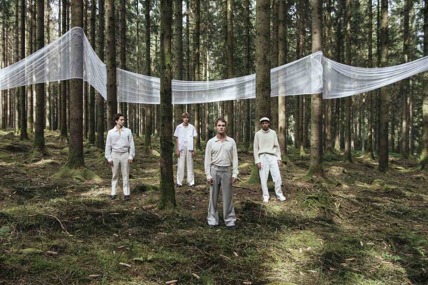The band standing among tall pine trees with lengths of white fabric strung between the trunks
